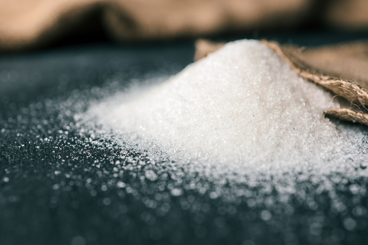 A small pile of white granulated sugar spills from a burlap sack onto a dark surface, with sugar crystals scattered around.