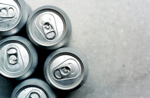 Five silver soda cans viewed from above are grouped together on the left side of a light grey textured surface, with condensation visible on the cans. The right side of the image is empty.