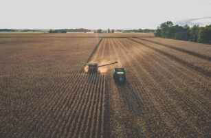 Aerial view of a combine harvester unloading crops into a tractor trailer in a large, golden cornfield at sunset, with trees and farmland in the background.