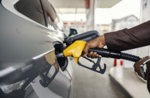 A person refuels a car at a gas station, holding a yellow fuel nozzle inserted into the vehicle’s fuel tank. The cars silver exterior and the station’s blurred background are visible.