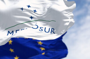 A white MERCOSUR flag with blue stars and a green line waves in the foreground, whilst the blue flag of the European Union with yellow stars is visible in the background against a partly cloudy sky.