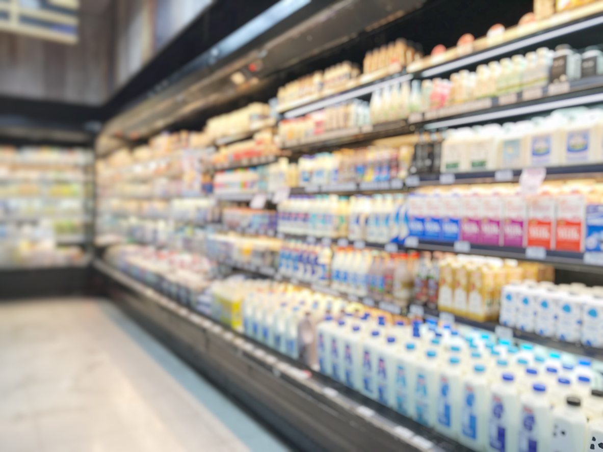 Blurred image of a supermarket dairy aisle with shelves stocked with various cartons and bottles of milk and other dairy products.