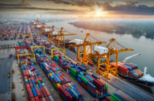 Aerial view of a busy shipping port at sunset, with colourful cargo containers stacked in rows, large yellow cranes, and a ship docked by the river. The city skyline is visible in the background.