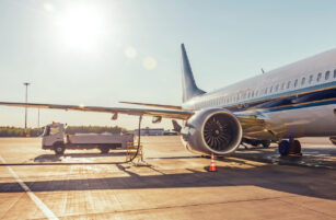 A commercial aeroplane is parked on the tarmac under bright sunlight, with a service lorry nearby and equipment connected to the aircraft. Trees line the background.