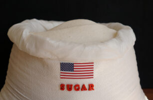 A large white sack filled with granulated sugar, labelled with red letters spelling SUGAR and displaying a small American flag on the front, placed against a dark background.