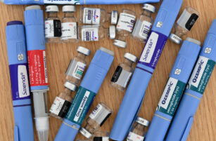 Several blue injector pens labelled Saxenda and Ozempic are scattered on a wooden surface among small clear vials with prescription labels, indicating various injectable medicines.