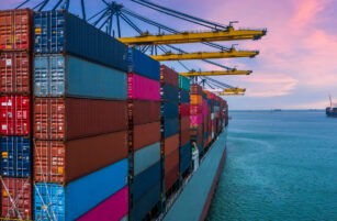 A large cargo ship loaded with stacked colourful shipping containers is docked at a port with yellow cranes under a pink and blue sunset sky, with more ships visible in the distance on the water.