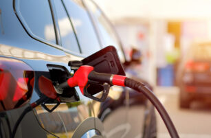 A close-up of a red petrol pump nozzle inserted into the petrol tank of a black car, with the fuel flap open at a petrol station. The background is blurred, showing other cars and the station area.