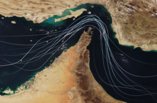 Satellite image of the Strait of Hormuz showing beige desert land and dark blue water, with white curved lines representing shipping routes crossing through the narrow sea passage.