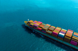A cargo ship loaded with colourful shipping containers sails through bright blue ocean water, leaving a wake behind as it moves. The image is taken from above, showing the ship and surrounding sea.