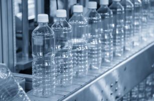 Clear plastic water bottles with white caps are lined up on an automated conveyor belt in a factory setting, suggesting a bottling or packaging process. The image has a cool blue tint.