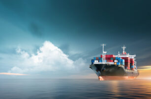 A large cargo ship loaded with colourful shipping containers sails on calm water under a dramatic sky with clouds and sunlight on the horizon.
