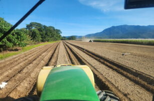 View from a green tractor driving along ploughed rows in a field, with trees and mountains in the background under a clear blue sky. Another vehicle is visible in the distance, raising dust.