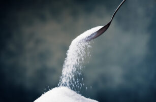 A spoon pours a stream of white granulated sugar onto a mound of sugar, set against a softly blurred dark background.
