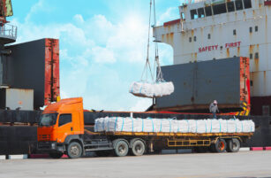 An orange lorry loaded with wrapped goods is parked beside a cargo ship. A crane lifts a bundle onto the lorry, while a worker stands on the lorry’s cargo. The sky is bright with clouds.