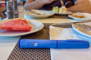 A blue insulin pen lies on a dining table set with plates of food, cutlery, a serviette, salt and pepper pots, and a slice of watermelon. A person in the background eats from a plate.