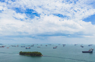 A small green island sits in blue ocean waters, surrounded by numerous cargo ships under a partly cloudy sky.