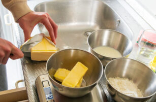 A person cuts a block of butter over a metal bowl near a sink. Other bowls containing milk, flour, and ingredients are on the worktop.