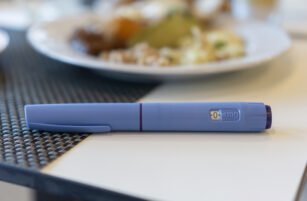 A blue medication pen labelled 0 mg lies on a white table, with blurred plates of food in the background.