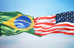 Two flags, one of Brazil (green, yellow, blue) and one of the United States (red, white, blue), wave together against a blue sky.