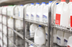 Shelves in a supermarket are stocked with multiple plastic gallon containers of milk, including varieties with red and blue caps. The focus is on the milk in the foreground, with more milk containers blurred in the background.