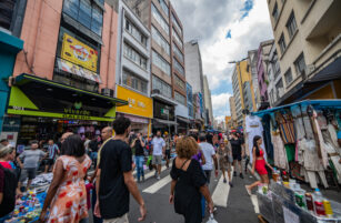 A busy urban street market with people shopping, street vendors, clothing stalls, and tall buildings on both sides under a partly cloudy sky.