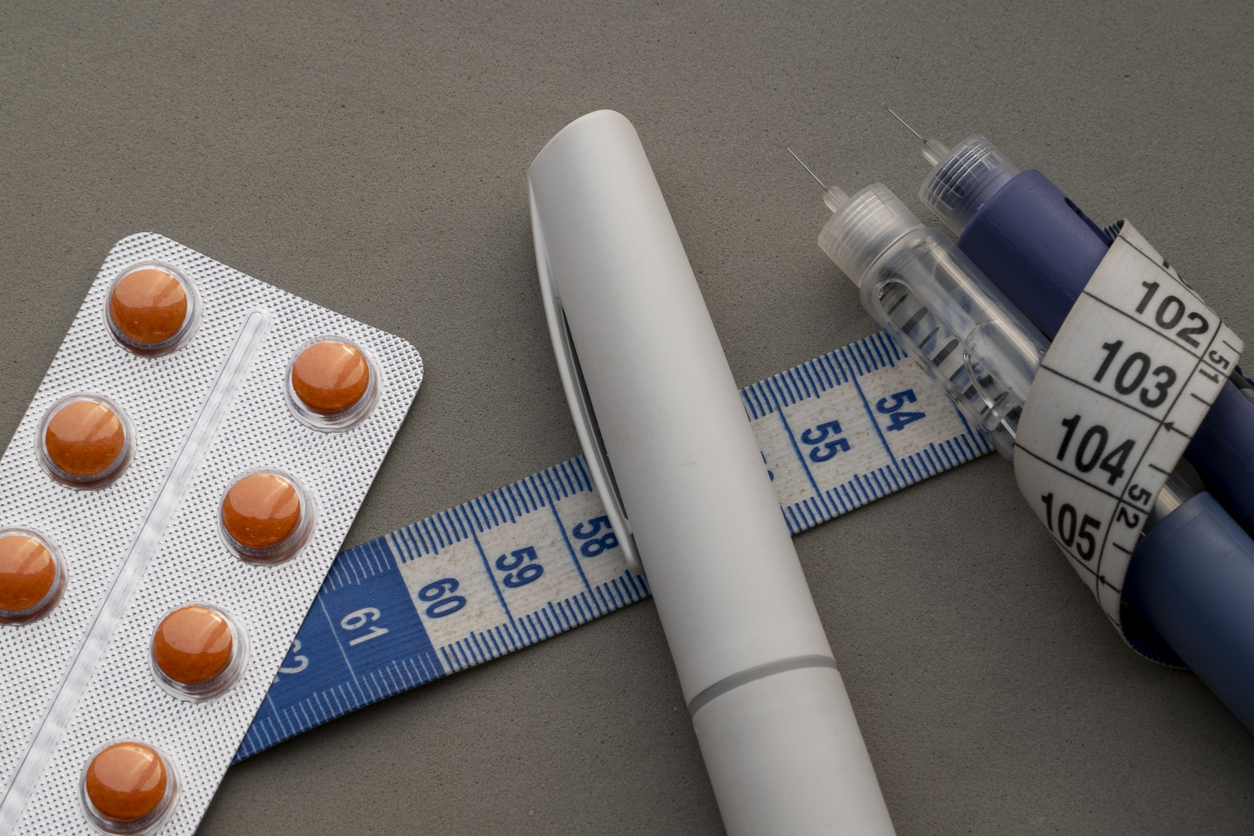 Tablets in a blister pack, two insulin pens, and a measuring tape are arranged on a grey background, symbolising diabetes management and health monitoring.
