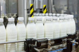 Plastic bottles filled with milk move along a conveyor belt in a factory setting, with machinery visible in the background.