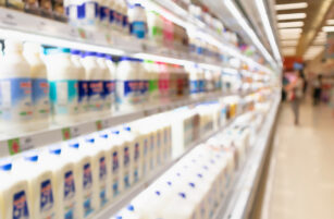 Blurred image of a supermarket aisle with shelves stocked with milk and dairy products, brightly lit under fluorescent lights.