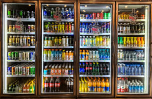 Three glass-door fridges display assorted bottled and canned drinks, including fizzy drinks, water, energy drinks, iced teas, and juices, all neatly arranged on shelves in a brightly lit convenience shop.