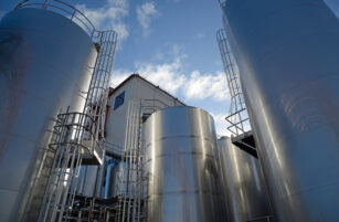 Large, shiny metal storage tanks with ladders surround an industrial building under a blue sky with clouds, viewed from a low angle.