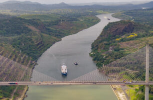 A large container ship travels through a wide canal flanked by green hills, with a modern cable-stayed bridge crossing above. Cars are visible on the bridge, and another ship is seen further down the canal.
