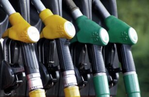 Four fuel pump nozzles in yellow, black, and green are lined up at a petrol station, ready for dispensing fuel. The background is slightly blurred.