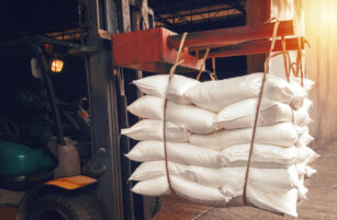 A forklift lifts a stack of white sacks, secured with straps, inside an industrial warehouse with sunlight streaming in from the right.