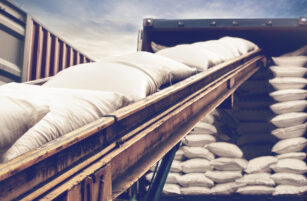 White sacks move up a conveyor belt into a shipping container filled with more sacks, under a partly cloudy sky.
