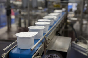 Several sealed white yoghurt pots move along a blue conveyor belt in a food processing factory, with industrial equipment blurred in the background.