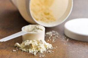 A plastic scoop filled with protein powder sits on a table, with more powder spilt around it. An open white container and its lid are in the background.