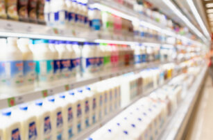 Blurred image of supermarket shelves stocked with various milk and dairy products in plastic containers, with bright lighting and colourful packaging.