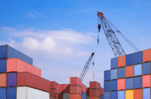 Stacks of colourful shipping containers with two cranes against a blue sky with scattered clouds, suggesting a busy container port or shipping yard.