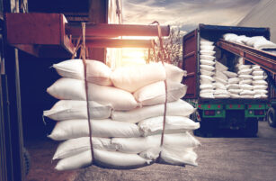 A forklift carries a large stack of white sacks towards a lorry bed filled with similar bags.
